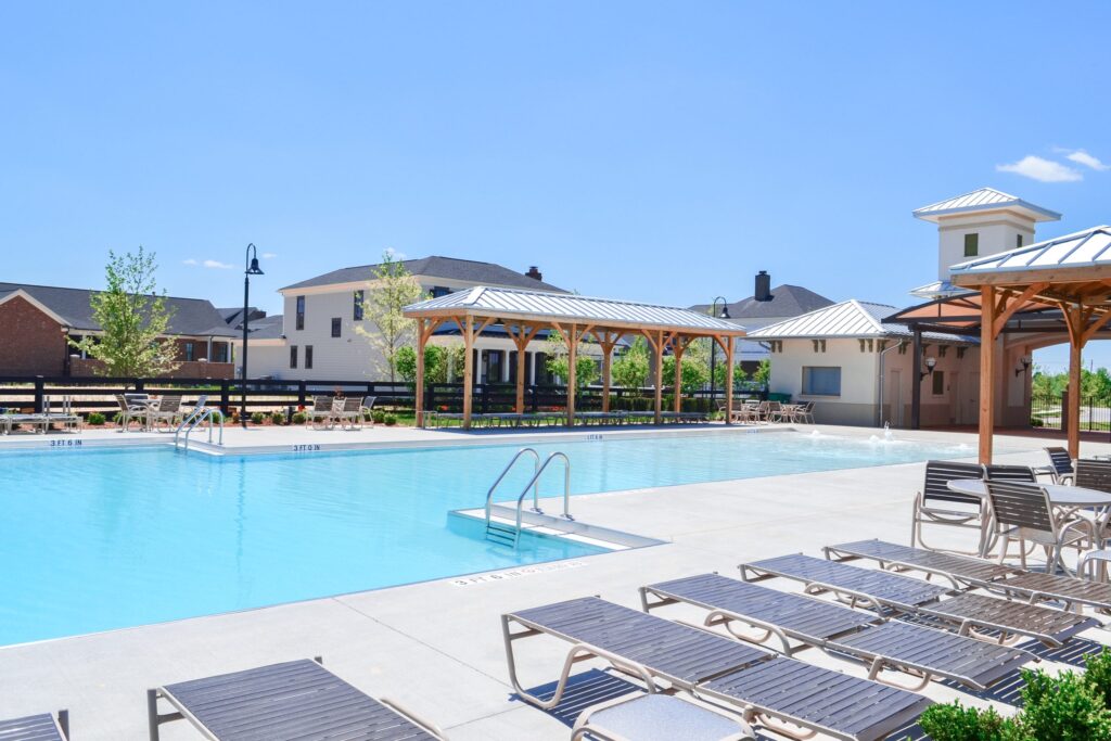 A clear blue swimming pool with adjacent lounge chairs under a clear sky. Covered seating areas are visible in the background, along with residential buildings.