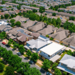 Aerial view of a suburban neighborhood with rows of houses, tree-lined streets, parked cars, and a green park area on the left.