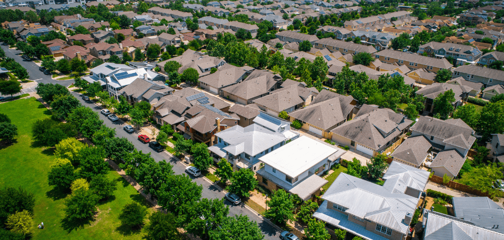 Aerial view of a suburban neighborhood with rows of houses, tree-lined streets, parked cars, and a green park area on the left.